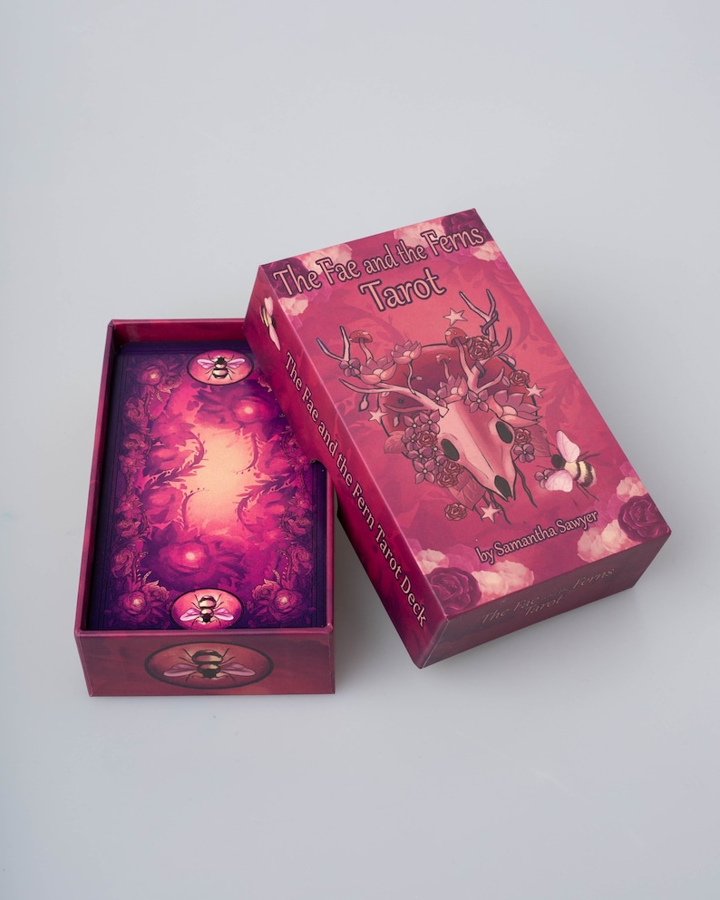 May include: A closed box of The Fae and the Ferns Tarot deck. The box is red with a pink and purple illustration of a deer skull with flowers and leaves. The text "The Fae and the Ferns Tarot" is printed in white on the box. The text "The Fae and the Fern Tarot Deck" is printed in white on the side of the box. The text "by Samantha Sawyer" is printed in white on the bottom of the box.