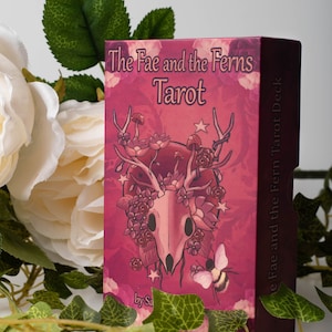 May include: A pink box with a skull and floral design. The box is for a tarot deck called "The Fae and the Ferns Tarot Deck".
