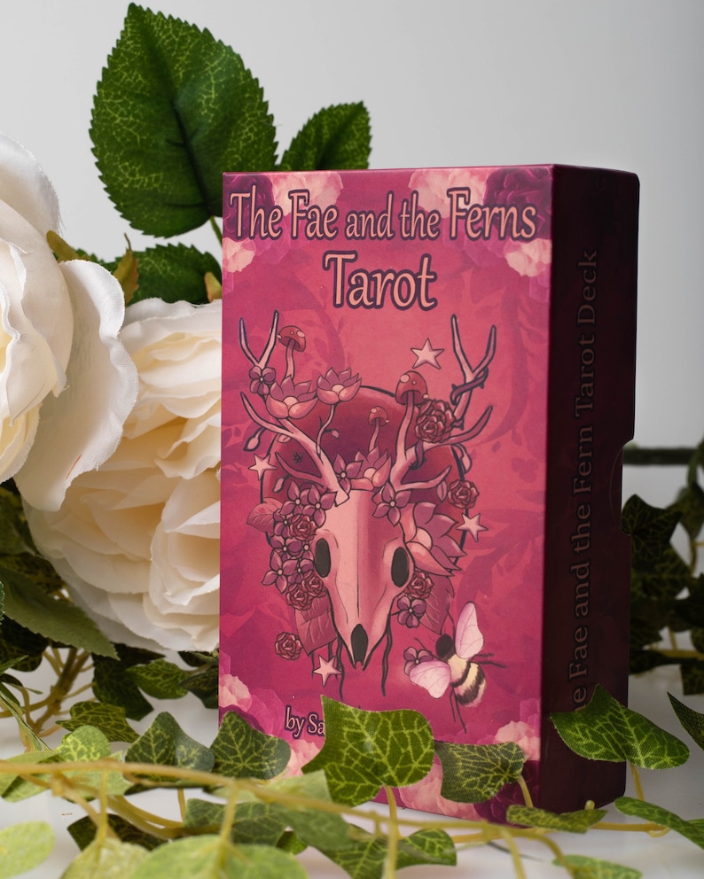 May include: A pink box with a skull and floral design. The box is for a tarot deck called "The Fae and the Ferns Tarot Deck".