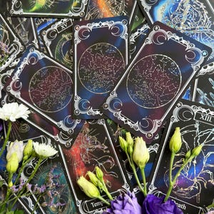May include: A collection of tarot cards featuring celestial and constellation designs with dark blue, purple, and black backgrounds. The cards display intricate white line art, including the labels The Hermit, Temperance, and Death, alongside white and purple flowers.