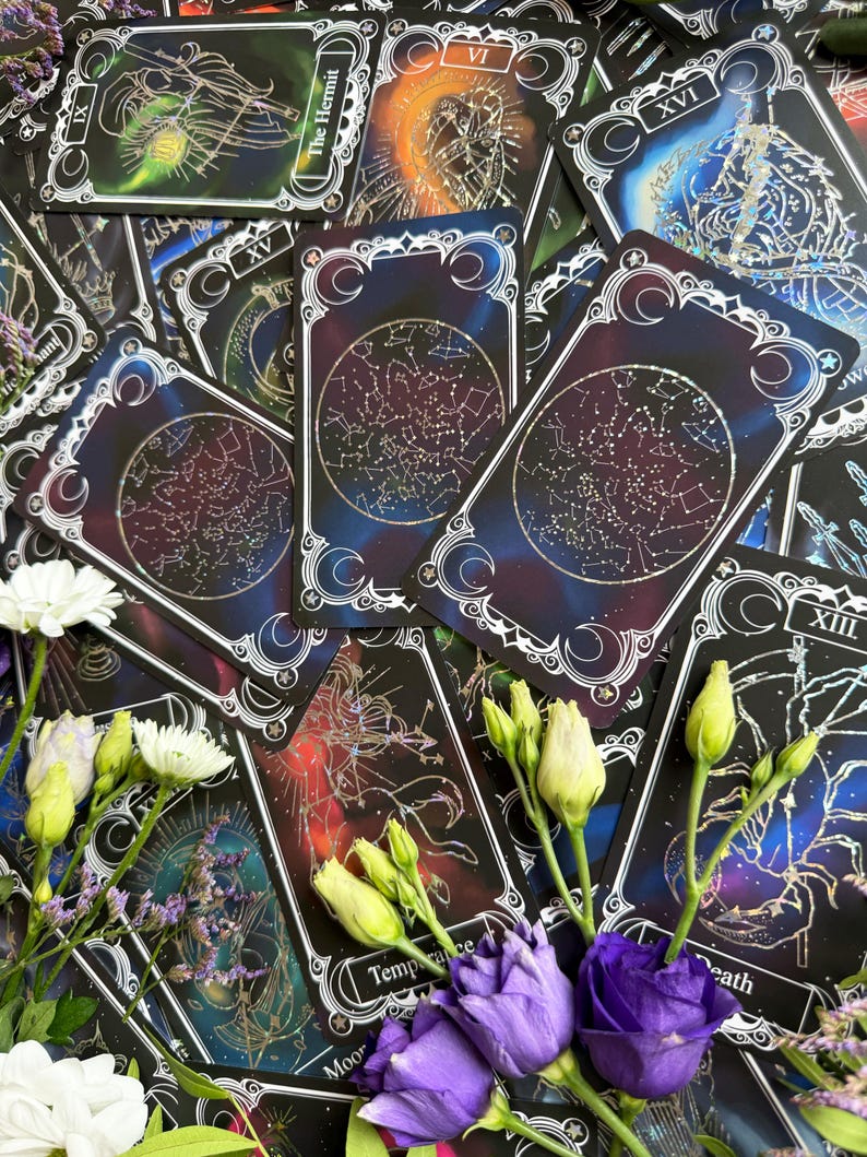 May include: A collection of tarot cards featuring celestial and constellation designs with dark blue, purple, and black backgrounds. The cards display intricate white line art, including the labels The Hermit, Temperance, and Death, alongside white and purple flowers.