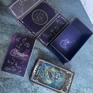 May include: A Stargazer Tarot deck set featuring a dark blue box with gold constellation patterns, an intuitive guide booklet with the text Stargazer Tarot Sam Sawyer, and a single card displaying The Moon with the Roman numeral XVIII.