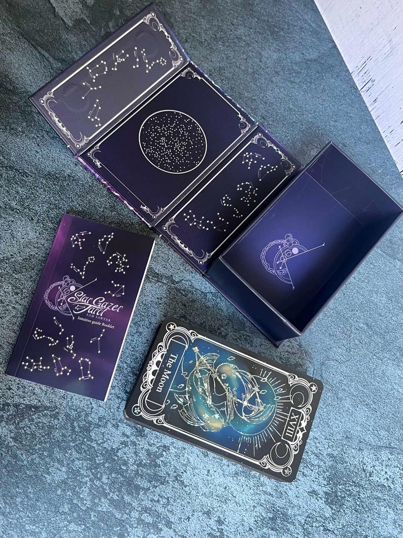 May include: A Stargazer Tarot deck set featuring a dark blue box with gold constellation patterns, an intuitive guide booklet with the text Stargazer Tarot Sam Sawyer, and a single card displaying The Moon with the Roman numeral XVIII.
