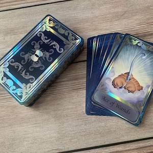 May include: A deck of tarot cards with dark blue backs featuring silver holographic filigree borders. One card is fanned out, displaying a brown creature holding a silver wand amidst clouds, with the text Ace of Wands printed at the bottom.