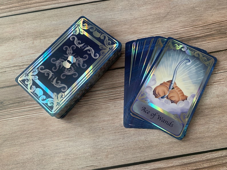 May include: A deck of tarot cards with dark blue backs featuring silver holographic filigree borders. One card is fanned out, displaying a brown creature holding a silver wand amidst clouds, with the text Ace of Wands printed at the bottom.