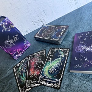 May include: A Star Gazer Tarot deck set featuring a purple and blue celestial-themed storage box, a stack of cards with constellation backs, three individual tarot cards including The High Priestess, and an intuitive guide booklet.