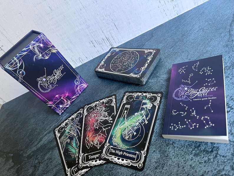 May include: A Star Gazer Tarot deck set featuring a purple and blue celestial-themed storage box, a stack of cards with constellation backs, three individual tarot cards including The High Priestess, and an intuitive guide booklet.