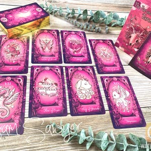 May include: The Fae and the Ferns Tarot deck featuring vibrant pink and purple illustrated cards with gold edges. The set includes a box with a skull and floral design, a guidebook, and several individual cards displaying whimsical creatures.