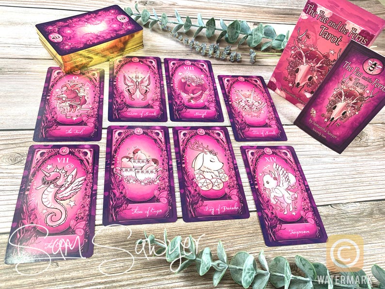 May include: The Fae and the Ferns Tarot deck featuring vibrant pink and purple illustrated cards with gold edges. The set includes a box with a skull and floral design, a guidebook, and several individual cards displaying whimsical creatures.