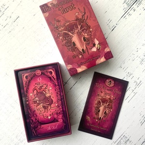 May include: A tarot deck box with a pink and purple design featuring a skull with antlers and flowers. The box has the text "The Fae and the Ferns Tarot" and "by Samantha Sawyer".