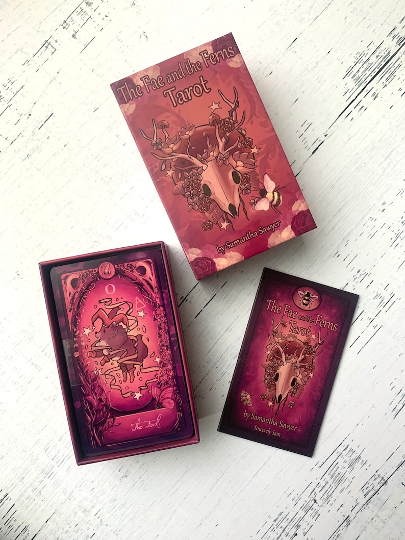 May include: A tarot deck box with a pink and purple design featuring a skull with antlers and flowers. The box has the text "The Fae and the Ferns Tarot" and "by Samantha Sawyer".