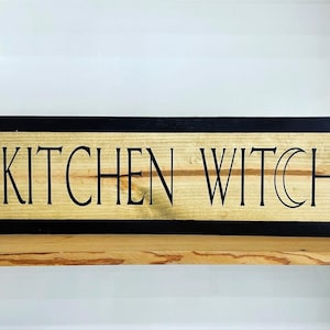May include: A wooden sign with a black frame and the text "KITCHEN WITCH" in black lettering.