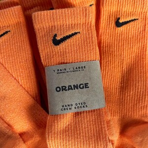 Pode incluir: Um par de meias laranja tingidas &agrave; m&atilde;o com um logotipo Nike preto em forma de swoosh. As meias s&atilde;o etiquetadas como "1 Pair - Large, Womens 10-13 Mens 8-12, Orange, Hand Dyed Crew Socks".