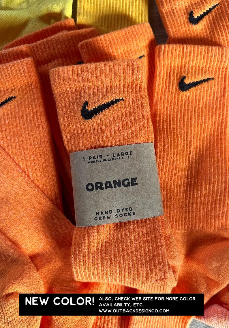 Pode incluir: Um par de meias laranja tingidas &agrave; m&atilde;o com um logotipo Nike preto em forma de swoosh. As meias s&atilde;o etiquetadas como "1 Pair - Large, Womens 10-13 Mens 8-12, Orange, Hand Dyed Crew Socks".