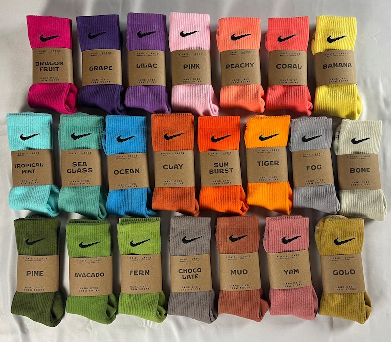 Pode incluir: Uma cole&ccedil;&atilde;o de 24 pares de meias de cano alto tingidas &agrave; m&atilde;o com diferentes cores e padr&otilde;es. Cada par tem uma etiqueta com o nome da cor e uma descri&ccedil;&atilde;o do processo de tingimento. As meias est&atilde;o dispostas em quatro filas de seis pares cada.