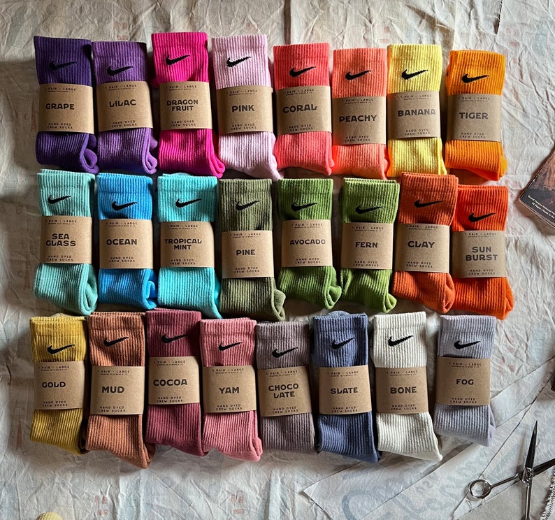 Pode incluir: Uma cole&ccedil;&atilde;o de 30 pares de meias tingidas &agrave; m&atilde;o em v&aacute;rias cores, incluindo roxo, rosa, verde, azul, laranja, amarelo e marrom. Cada par tem uma cor diferente e uma pequena etiqueta com um nome, como "Uva", "Lil&aacute;s", "Fruta do Drag&atilde;o", "Rosa", "Coral", "P&ecirc;ssego", "Banana", "Tigre", "Vidro do Mar", "Oceano", "Menta Tropical", "Pinho", "Abacate", "Samambaia", "Argila", "Explos&atilde;o de Sol", "Ouro", "Lama", "Cacau", "Inhame", "Chocolate Tarde", "Ard&oacute;sia", "Osso" e "N&eacute;voa".