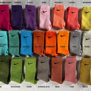 Pode incluir: Um conjunto de 24 meias coloridas com um logotipo da Nike. As meias est&atilde;o dispostas em quatro filas de seis meias cada. As meias s&atilde;o etiquetadas com nomes como "Fruta do Drag&atilde;o", "Uva", "Lil&aacute;s", "Rosa", "P&ecirc;ssego", "Coral", "Banana", "Menta Tropical", "Algas Marinhas", "Oceano", "Argila", "Explos&atilde;o de Sol", "Tigre", "N&eacute;voa", "Osso", "Pinho", "Abacate", "Samambaia", "Chocolate", "Lama", "Inhame" e "Ouro".