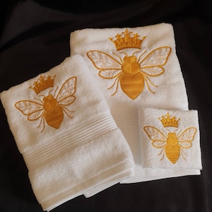 May include: Set of three white towels with embroidered gold bees and crowns. The towels are different sizes and are folded.