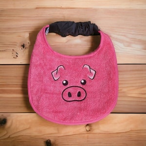 May include: Pink baby bib with a pig face design. The bib is made of soft, absorbent material and has a velcro closure.