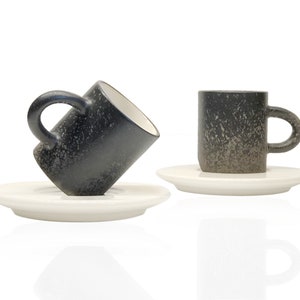 May include: Two black ceramic coffee cups with white saucers. The cups have a speckled, textured finish. One cup is tilted on its side.