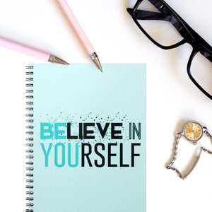 May include: A blue and white spiral bound notebook with the text "BELIEVE IN YOURSELF" in black letters. The notebook is on a white surface with two pink pens and a pair of black eyeglasses.