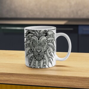 Może przedstawiać: Biały kubek ceramiczny z czarno-białym rysunkiem głowy lwa. Grzywa lwa jest ozdobiona skomplikowanymi wzorami.