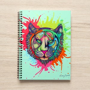 May include: A spiral bound notebook with a colorful illustration of a tiger's face on the front cover. The tiger is drawn in a detailed, intricate style with bright colors and a green background. The background also features colorful paint splatters.