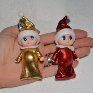 May include: Two small elf dolls with white fur hats and shiny fabric outfits. One doll is wearing a gold outfit and the other is wearing a red outfit.