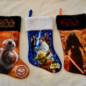 May include: Three Star Wars themed Christmas stockings. The first stocking features BB-8 from Star Wars: The Force Awakens. The second stocking features Luke Skywalker, Han Solo, Yoda, and Chewbacca from Star Wars: A New Hope. The third stocking features Kylo Ren from Star Wars: The Force Awakens.