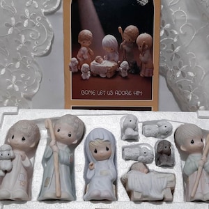 May include: A porcelain nativity set featuring figures of Mary, Joseph, shepherds, and animals. The figures are in pastel colors and are arranged in a white tray. A card with the text "COME LET US ADORE HIM" is in the background.