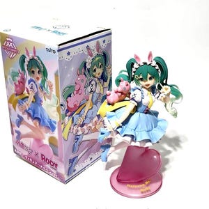 May include: A Hatsune Miku figurine with green hair and a blue dress, holding a pink pig-like creature. The figurine is on a pink heart-shaped base. The box features the same character in a similar pose.