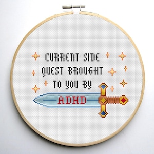 Puede incluir: Un aro de bordado blanco con un diseño de punto de cruz que presenta una espada con una hoja azul y un mango dorado. El texto "CURRENT SIDE QUEST BROUGHT TO YOU BY ADHD" está bordado en hilo negro.