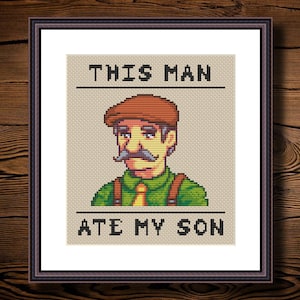 Puede incluir: Imagen enmarcada de punto de cruz que representa a un hombre de dibujos animados con bigote y gorra marrón. El texto encima de la imagen dice "THIS MAN" y el texto debajo dice "ATE MY SON". La obra está enmarcada en un marco marrón oscuro.