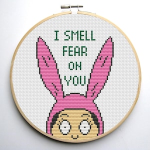 Puede incluir: Un bastidor de punto de cruz con un personaje de dibujos animados con un gorro de conejito rosa. El texto verde "I SMELL FEAR ON YOU" está bordado sobre la cara del personaje. El bastidor es de madera.