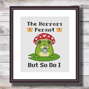 May include: Framed cross-stitch art. A green frog with a red mushroom hat and a cup is the central image. Text reads "The Horrors Persist But So Do I" with butterfly motifs. The artwork is set against a white backdrop within a dark brown frame.