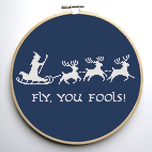 Puede incluir: Bordado de punto de cruz en un aro de madera. El diseño presenta a un Papá Noel blanco en un trineo tirado por renos, con el texto "fly, you fools!" sobre un fondo azul marino.