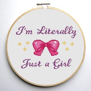 May include: Cross stitch embroidery hoop with the text "I'm Literally Just a Girl" in purple thread. A pink bow with yellow stars is stitched above the text.