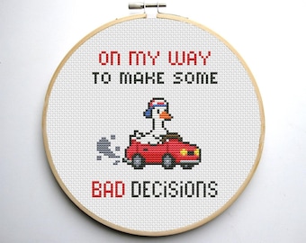 May include: Cross-stitch embroidery hoop with a white background and a red car with a goose wearing a blue cap driving. The text reads "On my way to make some bad decisions".