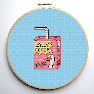 Może przedstawiać: Haft krzyżykowy przedstawiający różowe pudełko z sokiem z napisem "SILLY GOOSE JUICE 100%" i ilustracją gęsi. Projekt umieszczono na jasnoniebieskim tle w drewnianym tamborku.