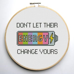 May include: A white embroidery hoop with a cross-stitch design that reads "Don't let their energy change yours". The design features a battery with the word "energy" in rainbow colors.