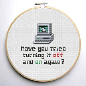 Op de afbeelding: Een witte kruissteek borduurring met een gepixelde afbeelding van een computer en de tekst "Heb je geprobeerd om het uit en weer aan te zetten?" in zwart en rood.