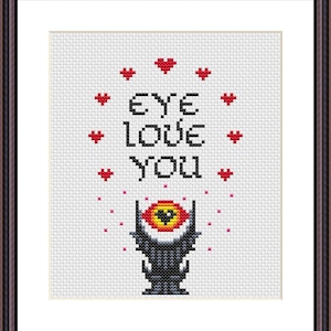 May include: Cross-stitch artwork in a brown frame. The design features the text "EYE LOVE YOU" above a stylized eye with a yellow iris and red accents. Red hearts and pink dots surround the text.