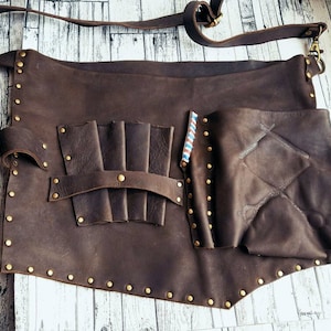 Puede incluir: Un delantal de cuero marrón con múltiples bolsillos y una correa. El delantal está decorado con tachuelas de latón y tiene una solapa en el lado derecho.