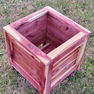 May include: A square wooden planter box made of cedar wood. The planter has a natural reddish-brown color with visible wood grain. The planter is empty and ready for planting.