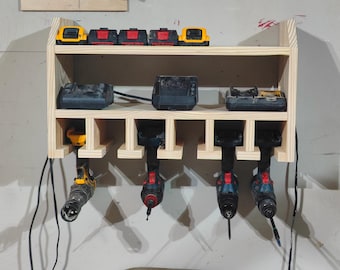 May include: A wooden wall-mounted storage rack with multiple compartments for holding power tools and batteries. The rack has a top shelf for storing batteries and a lower shelf with slots for holding power tools. The rack is made of unfinished wood and has a natural wood color.