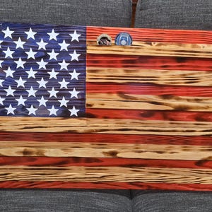 May include: Wooden American flag with a blue field of white stars and red and natural wood stripes. The flag has a distressed, rustic appearance. Two small coins are placed on the flag.