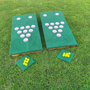 May include: A green and brown outdoor game with two boards, each with a triangle of white cups. There are yellow golf balls on the green grass.