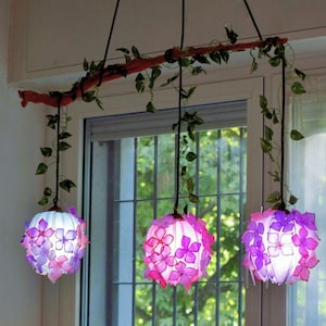 Op de afbeelding: Drie bolvormige hanglampen opgehangen aan een horizontale houten tak versierd met groene kunstmatige klimop. Elke lamp heeft een doorschijnende witte basis bedekt met gelaagde paarse en roze bloemvormige blaadjes die oplichten.