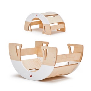 Puede incluir: Un juguete de madera para mecer con un acabado blanco y madera natural. El juguete tiene una forma curva y dos asas a los lados. El juguete está etiquetado con "SOY YO" en el lateral.