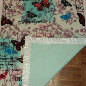 May include: A soft, rectangular blanket with a butterfly and floral pattern. The blanket features butterflies in shades of red, blue, and black, with floral and script accents. The edges are finished with a white satin trim. The reverse side is a solid light green.